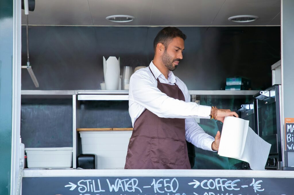 Young bearded waiter in uniform and apron standing at modern food truck counter with paper towel