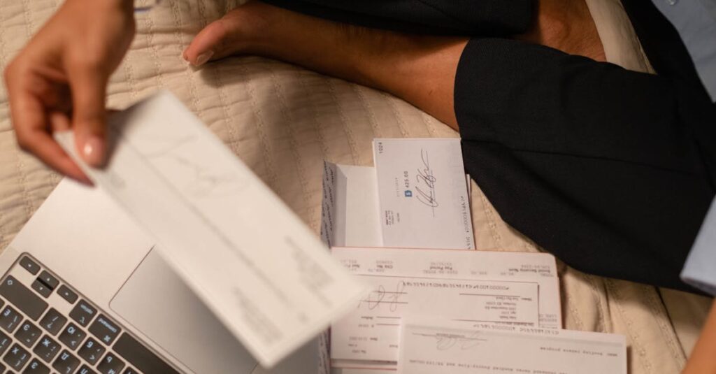 An adult organizing cheques and using a laptop indoors for financial tasks.
